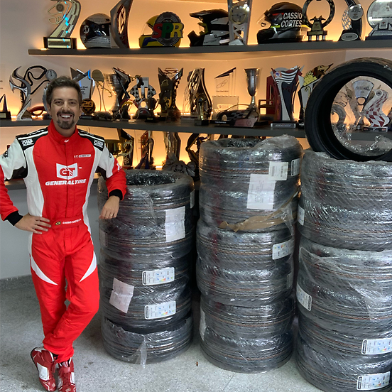 A imagem mostra Cassio Cortes com macacão de corrida vermelho e branco da General Tire, posando ao lado de várias pilhas de pneus embalados. Ao fundo, uma estante iluminada exibe troféus e capacetes, sugerindo um ambiente de corrida ou oficina. Ele sorri e apoia uma mão em uma das pilhas de pneus, transmitindo uma sensação de conquista e preparação para uma competição ou evento automobilístico.