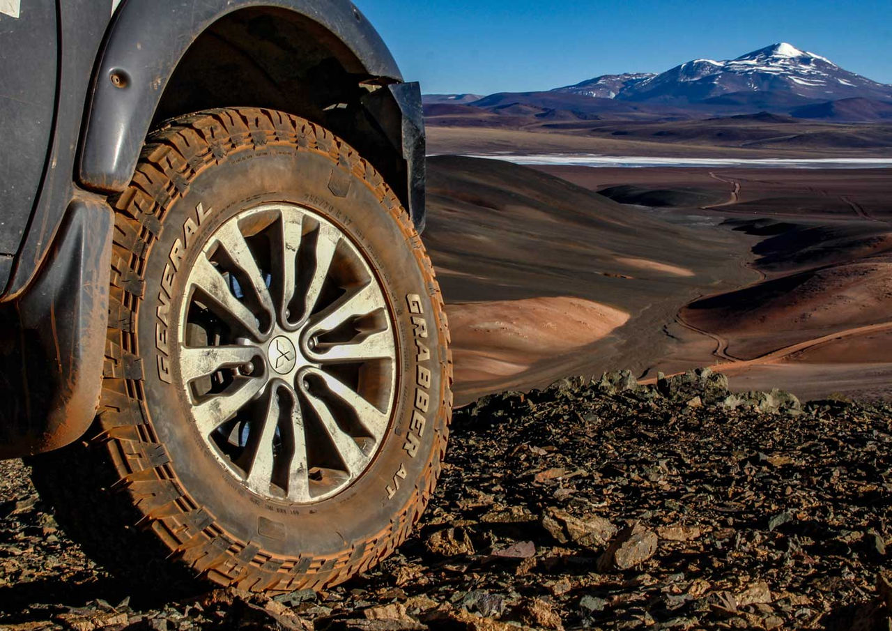 A imagem mostra um Grabber A/Tx da General Tire posicionado em um pico da Cordilheira dos Andes na Argentina e no fundo é possível ver outro pico com o cume cheio de neve. O terreno é bem pedregoso e agressivo, mas o Grabber A/Tx possui robustez para ele.