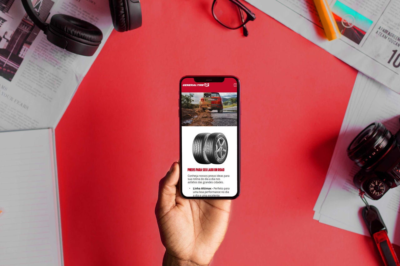 Imagem - Notícias sobre a Marca General Tire A imagem mostra uma mão segurando um smartphone que exibe uma página da General Tire sobre pneus off-road. Ao fundo, há uma mesa com vários itens, como um fone de ouvido, um par de óculos, uma câmera, um caderno e folhas de papel. O fundo é vermelho e a mesa está desorganizada.