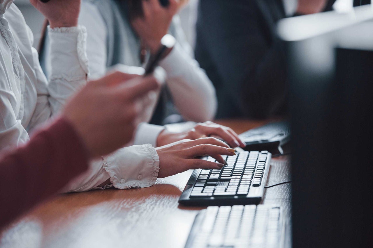 Imagem - Contato com a marca General Tire Na imagem, várias pessoas trabalham em um escritório, focadas em suas tarefas. As mãos de uma pessoa são vistas digitando em um teclado, enquanto outra segura um telefone. A foto destaca um ambiente profissional e dinâmico, onde a comunicação e o uso de computadores são fundamentais. As roupas formais sugerem um ambiente de trabalho corporativo.
