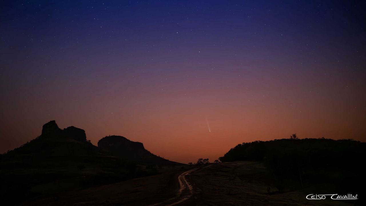 A imagem retrata uma paisagem noturna em uma atração turística chamada Três Pedras, em Bofete – município localizado no interior de São Paulo. Existe um céu estrelado e o cometa C/2023 A3 (Tsuchinschan-ATLAS) visível no horizonte, deixando um rastro luminoso no céu. O terreno é montanhoso, com duas grandes formações rochosas escuras em silhueta ao fundo. Um caminho de terra atravessa a imagem em direção ao horizonte. O céu apresenta uma transição de cores, indo do azul escuro no topo para tons alaranjados perto do horizonte, sugerindo o crepúsculo.