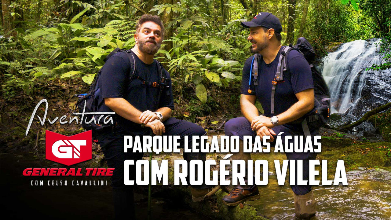 Celso Cavallini e Rogério Vilela estão sentados em um ambiente de mata atlântica, cercados por vegetação verde e uma pequena cachoeira ao fundo. Ambos usam roupas e mochilas de aventura. À esquerda, Rogério Vilela olha para o lado com expressão pensativa, enquanto à direita Celso Cavallini sorri para ele. O texto na imagem diz: "Aventura General Tire com Celso Cavallini - Parque Legado das Águas com Rogério Vilela". Ambiente transmite natureza e exploração.