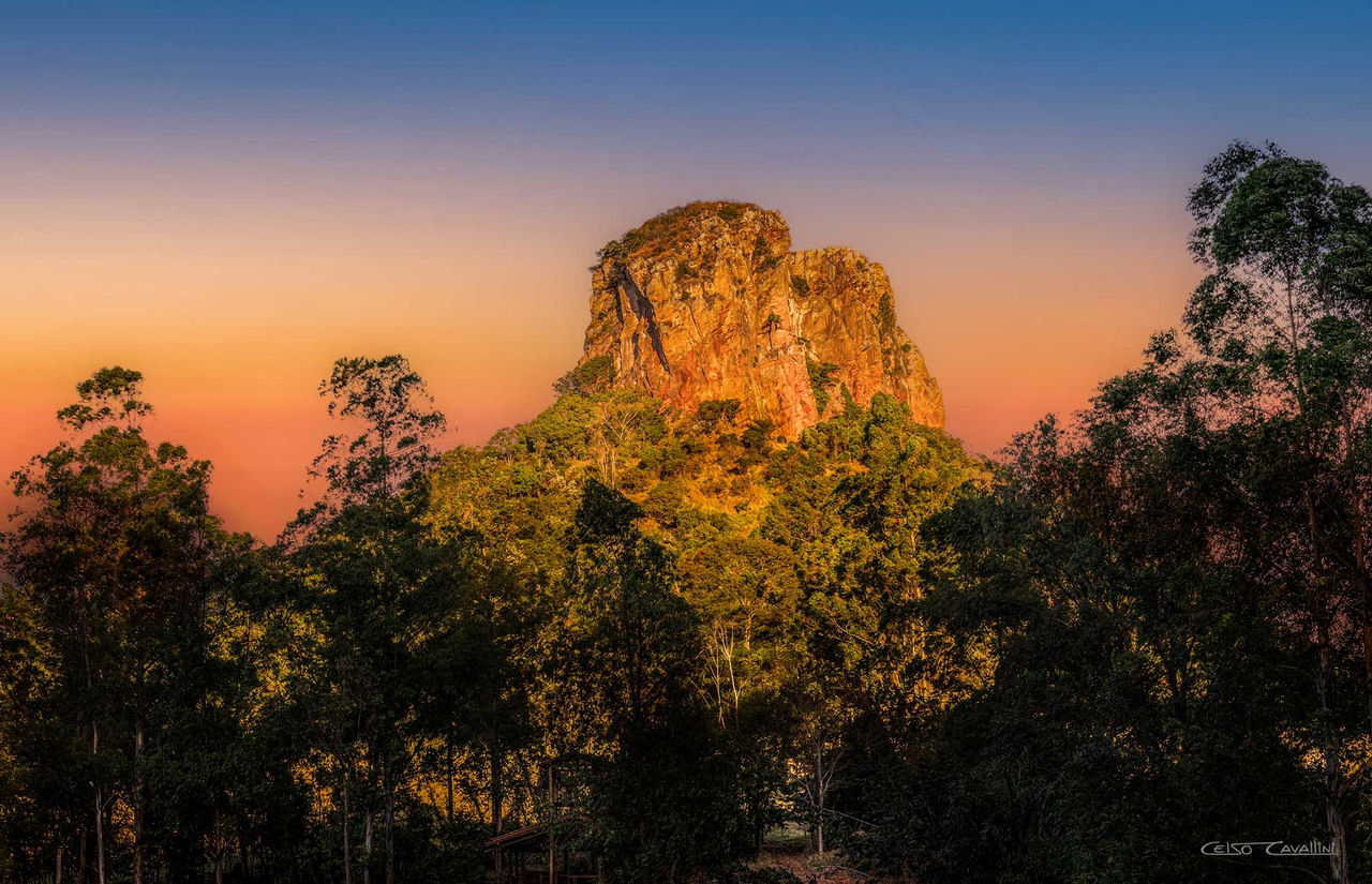 Imagem do Morro do Cuscuzeiro ao pôr do sol, com a luz dourada iluminando a formação rochosa e destacando suas cores avermelhadas. Árvores verdes cercam a base do morro, criando um contraste natural impressionante com o céu que gradua de laranja a azul. Uma pequena estrutura é visível no canto inferior direito, meio escondida pela vegetação.