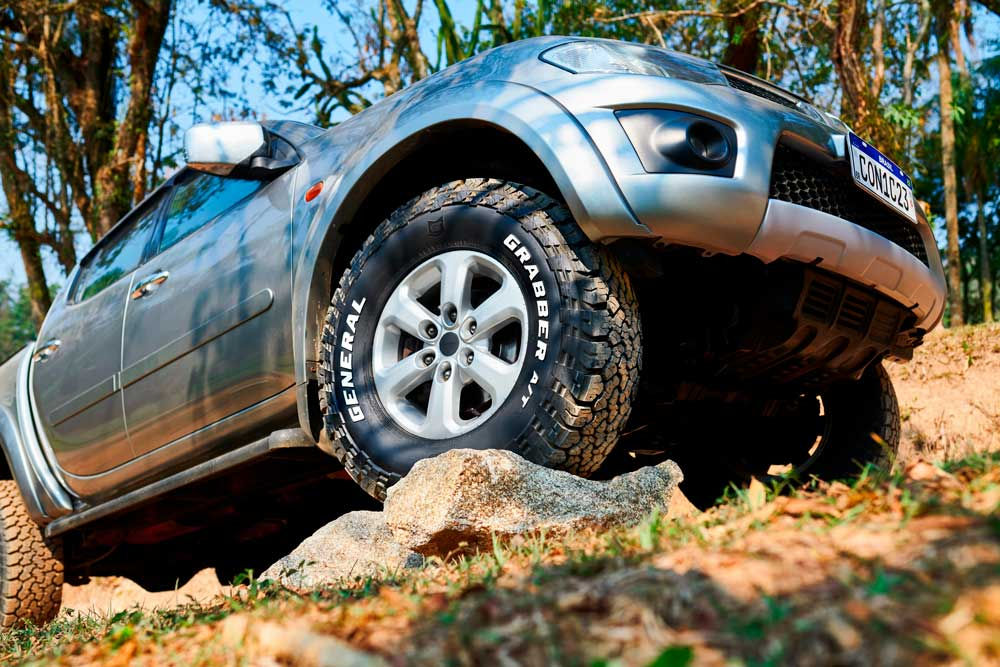 Um carro off-road cinza está subindo uma rocha em um ambiente natural com árvores ao fundo. O veículo está equipado com pneus General Tire Grabber A/Tx, projetados para terrenos mistos.