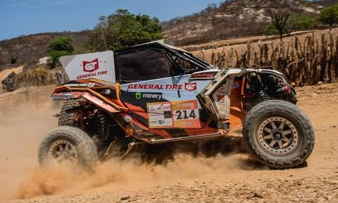 Um veículo de corrida de rally está em movimento, levantando poeira enquanto atravessa um terreno seco e acidentado. O carro é patrocinado pela General Tire, conforme indicado pelos logos na lataria. Atrás do veículo, há uma cerca de madeira e colinas cobertas de vegetação esparsa. O ambiente reflete uma típica paisagem de corrida off-road, com o carro projetado para resistir a condições extremas.