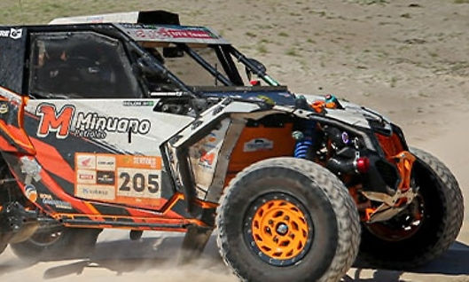 Um veículo de corrida de rally está em movimento, levantando poeira enquanto atravessa um terreno seco e acidentado. O carro é patrocinado pela General Tire, conforme indicado pelos logos na lataria. O ambiente reflete uma típica paisagem de corrida off-road, com o carro projetado para resistir a condições extremas.