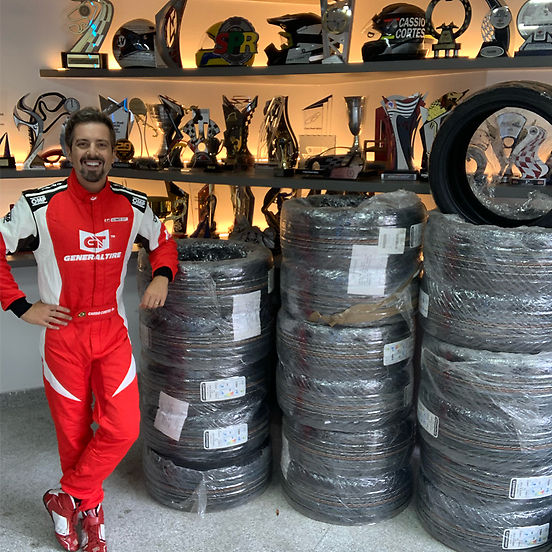 A imagem mostra Cassio Cortes com macacão de corrida vermelho e branco da General Tire, posando ao lado de várias pilhas de pneus embalados. Ao fundo, uma estante iluminada exibe troféus e capacetes, sugerindo um ambiente de corrida ou oficina. Ele sorri e apoia uma mão em uma das pilhas de pneus, transmitindo uma sensação de conquista e preparação para uma competição ou evento automobilístico.