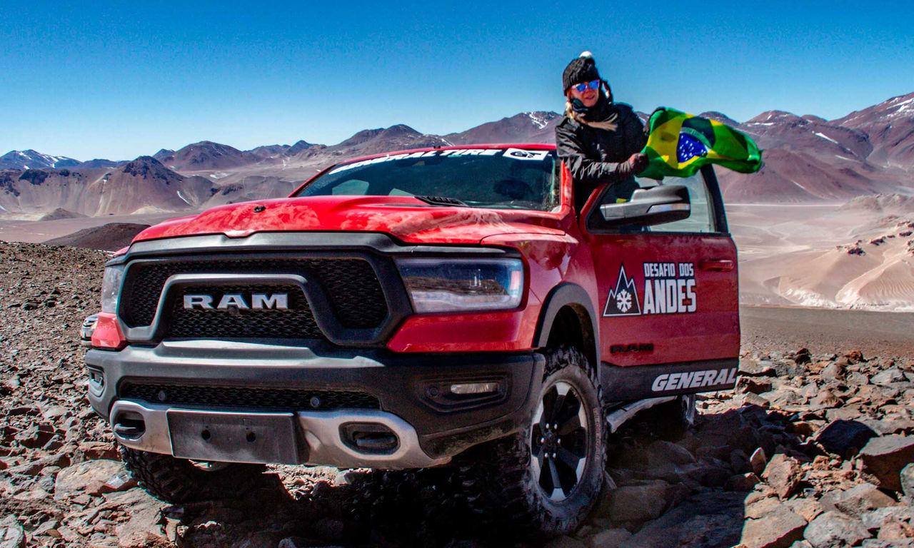 Imagem de uma pickup vermelha Dodge Ram na cordilheira dos andes com um terreno bem pedregoso. Posicionada a porta do veículo está a piloto Amanda Vasconcellos com a bandeira do Brasil na mão.